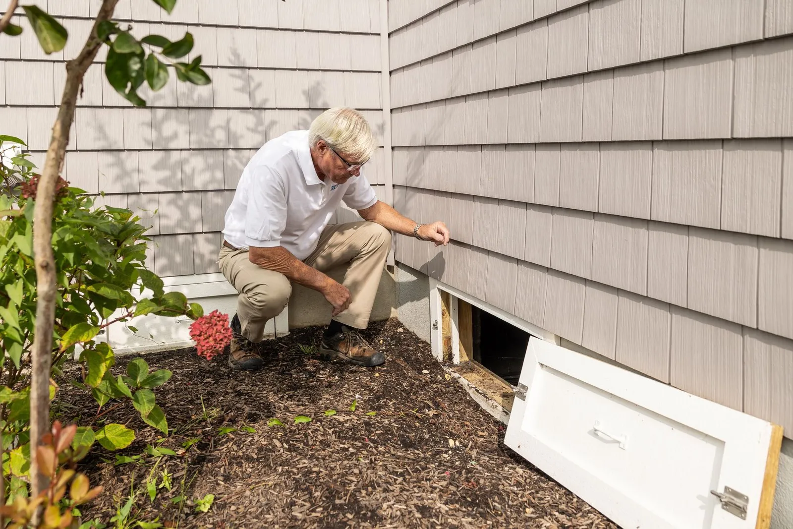 A home inspector kneeling at an open crawl space vent along the foundation of a house, checking for pest entry points.